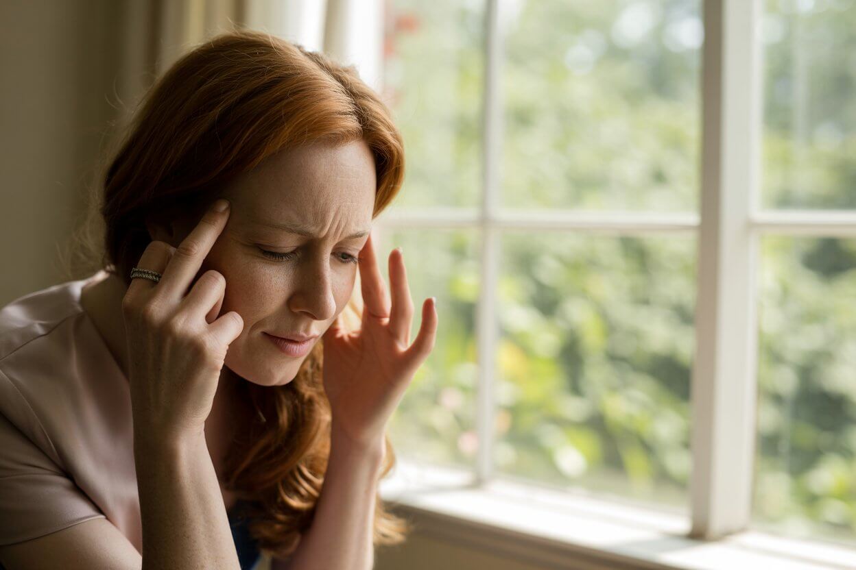 Woman with migraine pain holding her head near a sunny window.