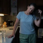 Woman pressing hand to cool countertop at night, lifting hair from neck during a hot flash.
