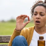 Tired young woman on a park bench yawning while holding up an iron capsule and a supplement bottle